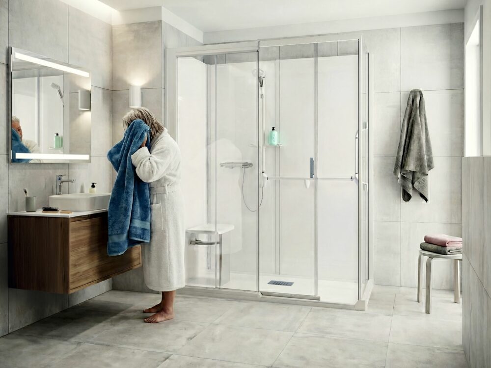 Ältere Frau im weißen Bademantel trocknet sich das Gesicht mit einem blauen Handtuch in einem modernen, barrierearmen Badezimmer von Kinedo mit bodengleicher Dusche und Glasabtrennung.